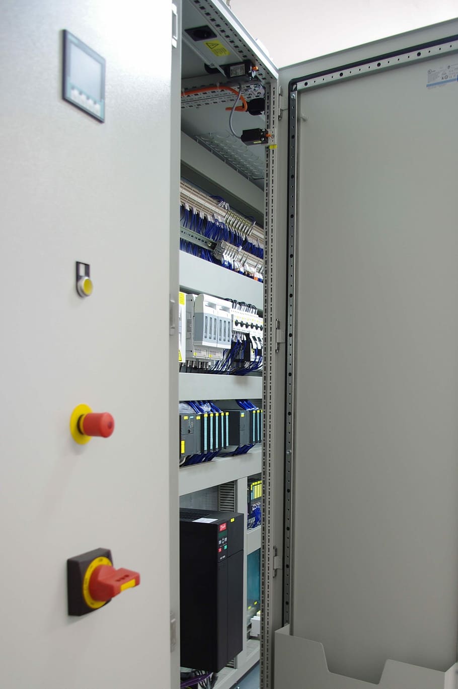 Cuadro de control industrial abierto con pantalla HMI, seta de emergencia, módulos de PLC y variador de frecuencia