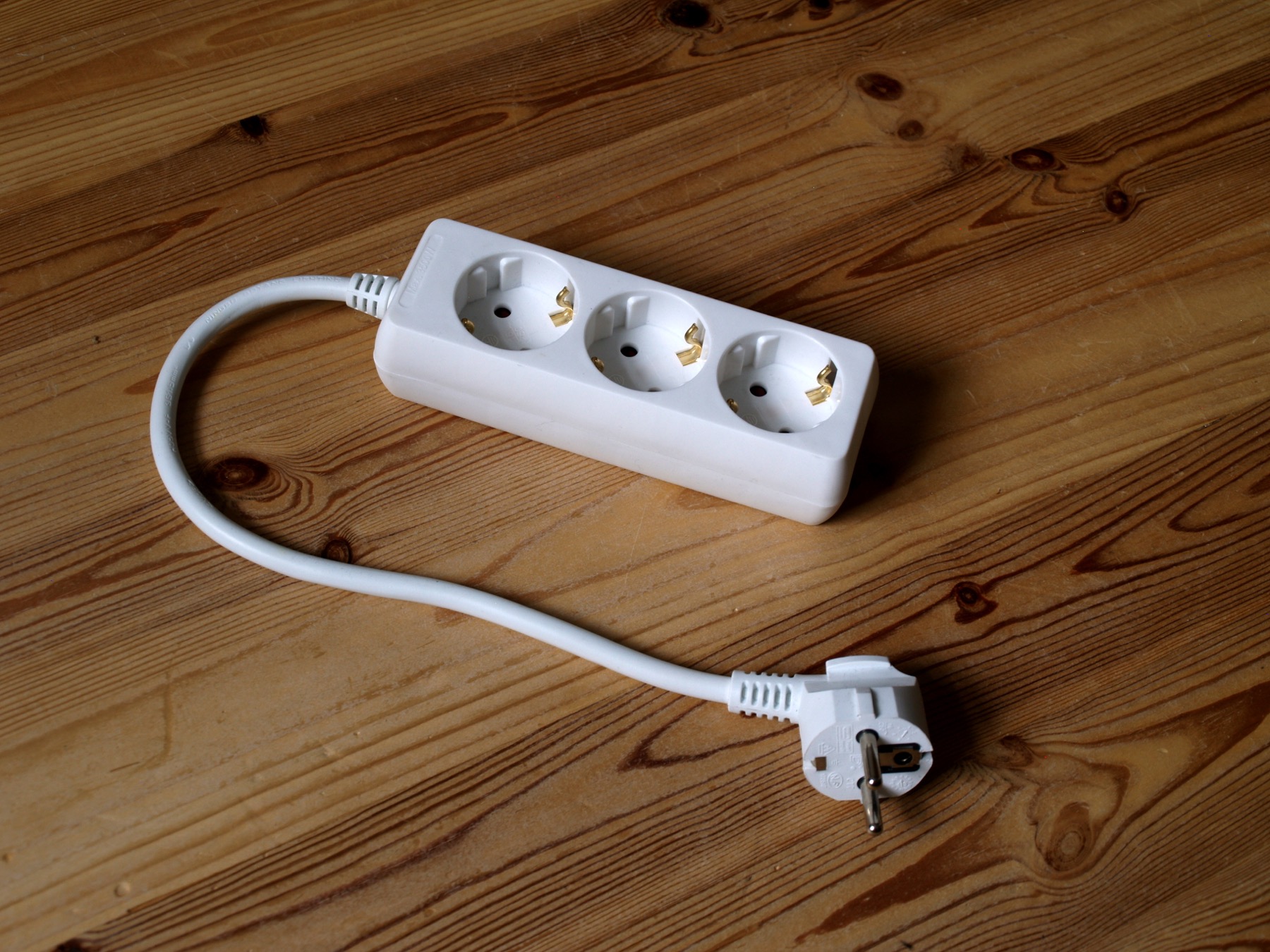 Regleta blanca de tres enchufes schuko con su cable y clavija sobre un suelo de madera