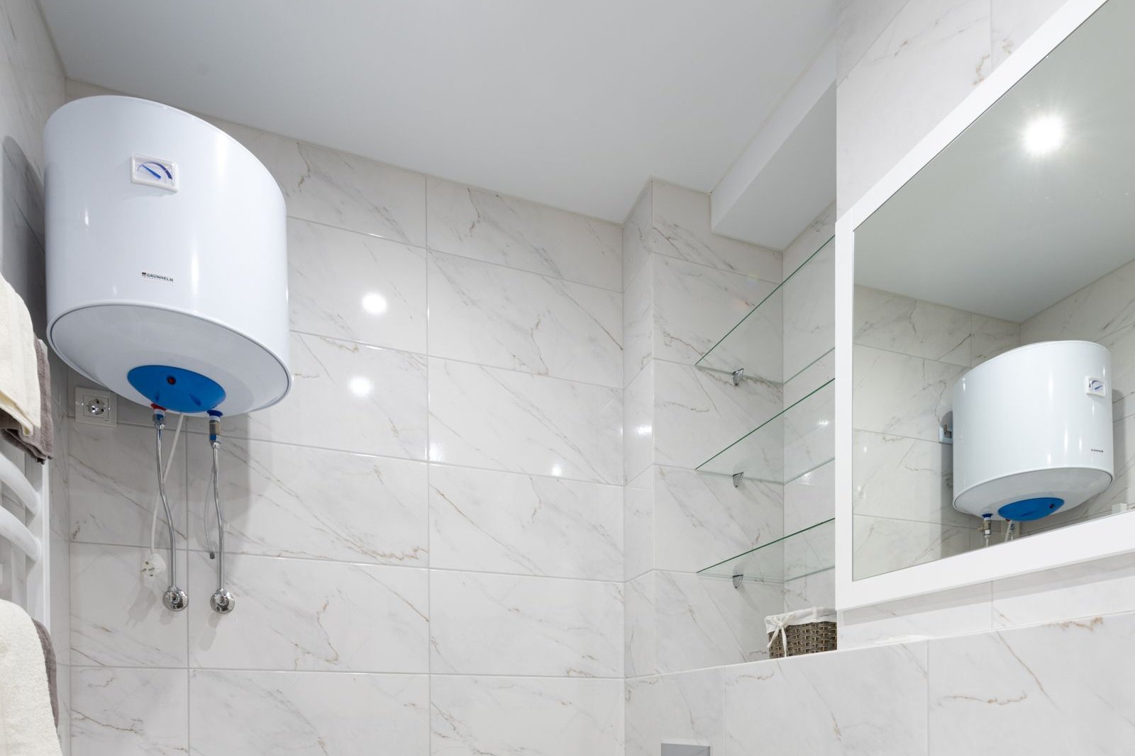 Termo eléctrico blanco instalado en la pared de un baño con azulejos de mármol y sus tomas de agua fría y caliente